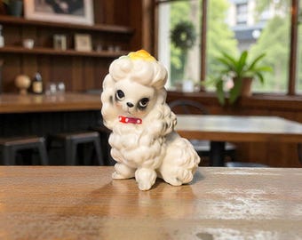 May include: A vintage white poodle figurine with a red collar. The poodle has a fluffy, textured coat, black eyes, and a yellow accent on its head. The figurine is sitting on a wooden surface.