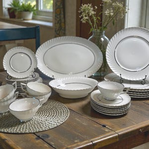 May include: A collection of white porcelain dinnerware with silver trim, including plates, bowls, cups, and saucers. The plates have a spiral design around the edges. The set is arranged on a wooden table.