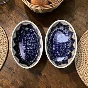 May include: Two oval serving bowls with a blue and white speckled design. The bowls have a white rim and a decorative blue border. The bowls are on a wooden table, with a basket of bread in the background.