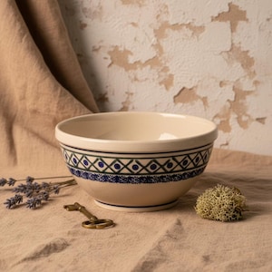 Może przedstawiać: Kremowa miska ceramiczna z ozdobnym pasem o geometrycznym wzorze ciemnoniebieskich i zielonych rombów oraz małych niebieskich kropek. Miska posiada gładką, zaokrągloną krawędź oraz lekko zwężającą się podstawę jako naczynie kuchenne.