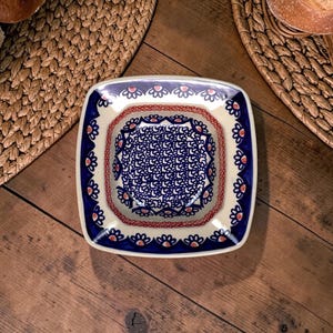 Könnte beinhalten: Quadratische Keramikservierschale mit blauem und weißem Blumenmuster. Die Schale hat einen weißen Rand mit blauen und roten Akzenten. Die Schale steht auf einem Holztisch, im Hintergrund Brot und ein geflochtener Platzdeckchen.