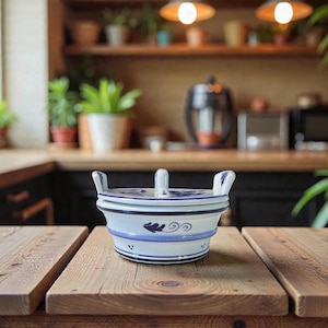 Könnte beinhalten: Eine weiß-blaue Keramikservierschale mit Deckel. Die Schale hat ein blaues Blumendesign und einen Griff auf dem Deckel. Die Schale steht auf einem Holztisch. Der Hintergrund zeigt eine Küche mit Pflanzen und Lichtern.