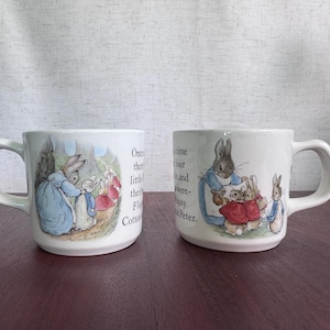 May include: Two white ceramic mugs featuring illustrations of Beatrix Potter's Peter Rabbit characters. The mugs display scenes of rabbits in blue jackets and red capes, accompanied by printed text from the story, including the phrase Once upon a time.