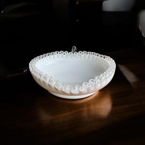 Puede incluir: Un cuenco blanco con borde con volantes y un asa transparente. El cuenco tiene un diseño ancho y poco profundo y se asienta sobre una pequeña base circular. El borde del cuenco presenta un delicado patrón ondulado. El cuenco está hecho de un material brillante y opaco.