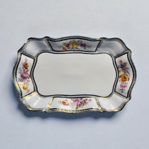 May include: A rectangular, white porcelain serving tray with a scalloped edge. The tray is decorated with floral patterns in shades of pink, yellow, and purple, and trimmed with gold and black accents. A decorative piece for serving.