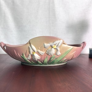 Può includere: Una ciotola ovale in ceramica con una finitura sfumata rosa e pesca. L'esterno è decorato con fiori di iris bianchi in rilievo con centri gialli e foglie verdi. La ciotola presenta piccole maniglie su ciascun lato.