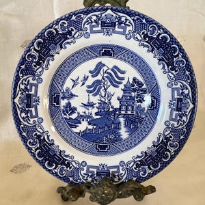 May include: A blue and white ceramic plate with a Willow pattern design. The plate features a central scene with a pagoda, bridge, and figures, surrounded by a decorative border. The plate is displayed on a decorative metal stand.