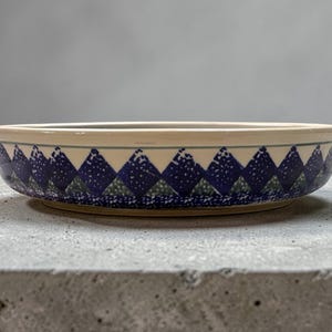 May include: A round ceramic bowl with a cream-colored rim and a blue and green geometric pattern around the sides. The pattern consists of diamond shapes with white speckles. The bowl sits on a gray surface.