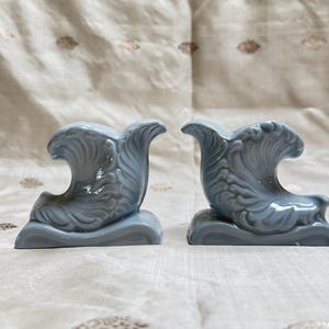 May include: Two matching, light blue ceramic wave sculptures. Each sculpture has a wave cresting over a base. The sculptures are displayed on a textured, off-white surface.