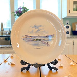 May include: A cream-colored decorative plate featuring a watercolor depiction of the San Francisco Bay, including the Golden Gate Bridge. The plate is displayed on a black metal stand on a wooden table.