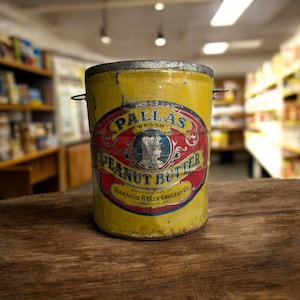 May include: Vintage yellow Pallas Peanut Butter tin with a red and blue label. The label features the text "Pallas Brand Peanut Butter" and a portrait. The tin has a metal lid and handles.