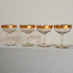May include: Four clear champagne coupe glasses with gold rims. The glasses have a classic design with a rounded bowl and a slender stem. The gold rim adds a touch of elegance to the glassware.