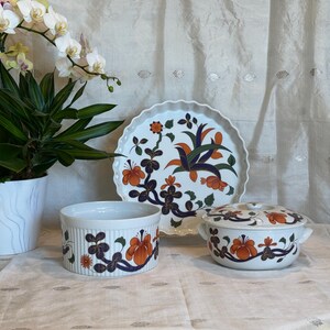 May include: Three white ceramic dishes with floral designs. The dishes feature orange, blue, and green floral patterns. The dishes are on a white tablecloth.