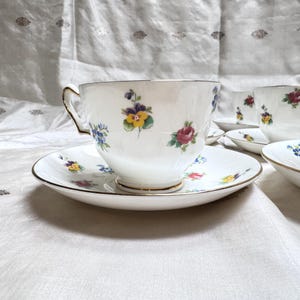 May include: White teacups and saucers with floral designs. The cups and saucers have a gold trim. The floral designs include purple, yellow, and red flowers. The teacups and saucers are arranged on a white surface.