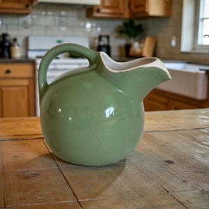 May include: A sage green ceramic pitcher with a rounded body and a curved handle. The pitcher has a spout and a white rim. It sits on a wooden table in a kitchen setting.