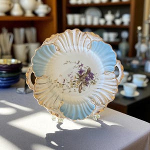 Puede incluir: Plato de porcelana antiguo con borde festoneado, detalles en azul claro y ribete dorado. El centro presenta un diseño floral con flores moradas y blancas. El plato tiene dos asas y está sobre un soporte transparente.
