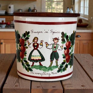 Peut inclure: Un pot de cuisine en céramique blanche avec un couvercle en bois foncé. Le pot est décoré d'une bande rouge, des mots "Sugar 'n' Spice" et "'n everything Nice", et d'un motif d'art populaire de personnes et de fraises.