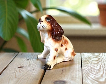 Peut inclure: Figurine en céramique représentant un chien, avec un pelage tacheté marron et blanc, des oreilles marron et un visage blanc avec des détails noirs. Le chien est assis, la tête levée. La figurine est posée sur une surface en bois.