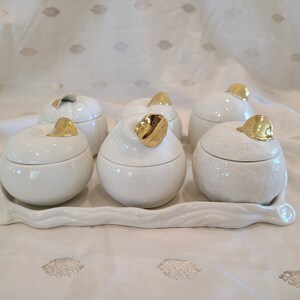 May include: Set of six white ceramic containers with gold accents. Each container has a different fruit design on the lid. The containers are arranged on a white tray.