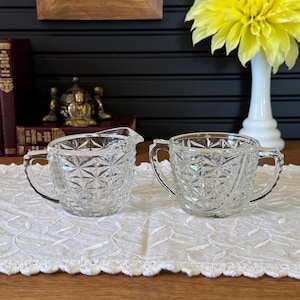 May include: A clear glass creamer and sugar bowl set with a diamond pattern. The set has handles and sits on a white lace tablecloth. A yellow flower in a white vase is in the background.