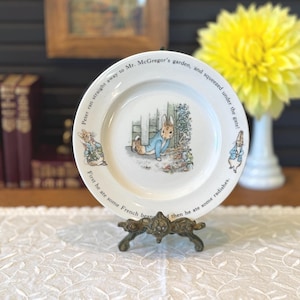 May include: A white ceramic plate featuring illustrations of Peter Rabbit in Mr. McGregor's garden. The plate has text around the rim, and is displayed on a decorative metal stand. A yellow flower and vase are in the background.