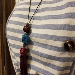 May include: A dark brown and blue beaded necklace with a maroon tassel. The necklace is on a striped blue and white background.
