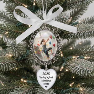 May include: Silver oval Christmas bauble with a photo of a person rock climbing. The bauble has a white ribbon and a heart-shaped charm with the text "2025 Hailey's first climbing comp". The bauble hangs on a Christmas tree.