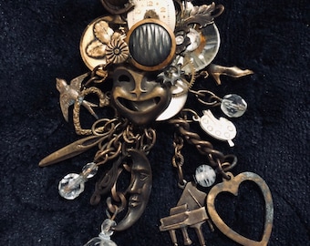 May include: A large, ornate, gold-toned pendant with a variety of charms, including a crescent moon, a heart, a piano, a bird, a clock face, and a mask. The pendant is made of metal and has a vintage aesthetic.