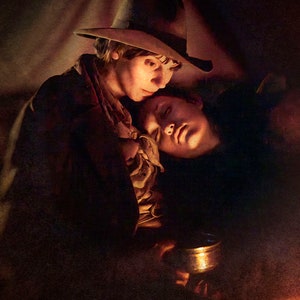 May include: A dimly lit image of two people in a tender embrace. One person, wearing a hat, looks down at the other, who rests their head on their shoulder. A golden chalice is held in the foreground, illuminated by a warm light.