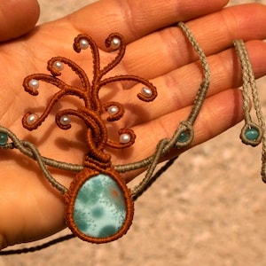 Puede incluir: Un collar con un colgante de árbol de la vida marrón y verde con una piedra turquesa y detalles de perlas blancas. El collar está hecho con cordón de macramé y tiene una combinación de colores marrón y verde.