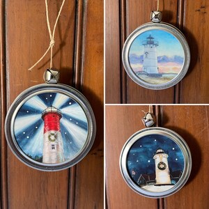 Op de afbeelding: Drie handgeschilderde vuurtorenversieringen met een zilveren metalen frame en een bel aan de bovenkant. De ornamenten tonen een vuurtoren met een rood-witte gestreepte toren, een witte vuurtoren met een blauwe lucht en zonsondergang, en een witte vuurtoren met een blauwe lucht en sterren.