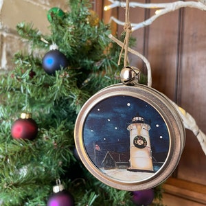 Puede incluir: Un adorno navideño plateado con una imagen pintada de un faro con una corona, sobre un fondo de cielo nocturno azul con estrellas blancas. El adorno está colgado de un árbol con otros adornos.