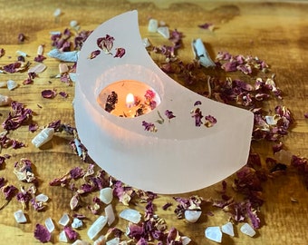 May include: A white selenite crescent moon candle holder with a small candle burning inside. The holder is surrounded by dried rose petals and small white crystals.