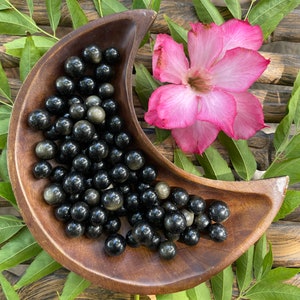 May include: A wooden bowl filled with black and gold gemstone spheres. The bowl is shaped like a crescent moon and is sitting on a woven surface with green leaves.