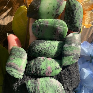 May include: A collection of seven green, black, and pink polished stones. The stones are smooth and have a mottled appearance.