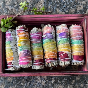 May include: A set of seven rainbow sage smudge sticks in a wooden box. The sticks are wrapped in colorful flower petals and tied with string.