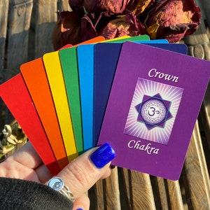 May include: A set of seven chakra cards with rainbow colors and a purple card with the word "Crown Chakra" and a symbol of the Om.
