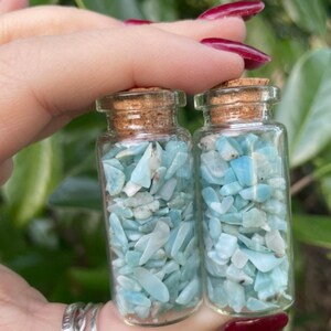 May include: Two small glass bottles filled with light blue and white aquamarine chips. The bottles have cork stoppers.