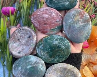 May include: Seven smooth, polished, oval-shaped stones in various shades of green, brown, and white. The stones are arranged in a semi-circle on a hand.