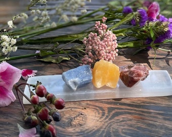 May include: Three crystals, a blue, a yellow, and a red, are arranged on a white selenite charging plate. The crystals are surrounded by flowers.