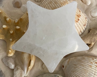 May include: A white, star-shaped selenite plate, surrounded by seashells and starfish on a sandy surface.