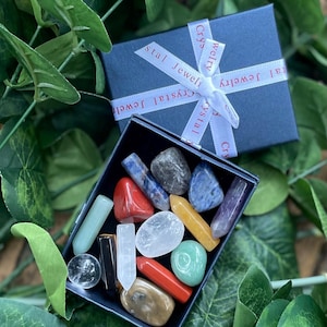 May include: A blue box with a white ribbon tied around it. The ribbon has the text "Crystal Jewelry" printed on it. The box is filled with a variety of colorful tumbled stones.
