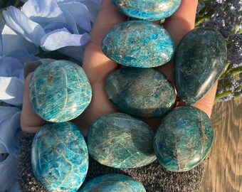 May include: A collection of polished, oval-shaped blue apatite stones. The stones vary in shades of teal and turquoise, with natural veining patterns. The stones are held in a hand, with blue flowers in the background.