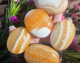 May include: Five polished, oval-shaped stones in shades of yellow and orange. Some stones have white accents or stripes. The stones are smooth and appear to be natural, with variations in color and pattern. The background includes flowers and wood.
