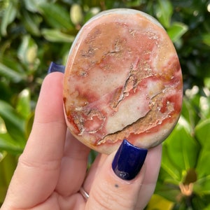 May include: A smooth, oval-shaped, polished stone with a unique pattern of red, brown, and white swirls and veins. The stone has a natural, earthy look.
