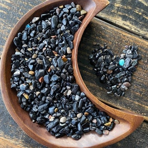 May include: A wooden crescent-shaped bowl filled with polished black stones. A heart shape made of similar stones sits beside the bowl. The stones have a glossy finish, with a few tan, gray, and blue stones mixed in.