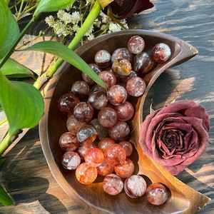 May include: A wooden bowl shaped like a crescent moon holds a collection of red and clear crystal spheres. The spheres have a cloudy, translucent appearance with hints of white and brown.