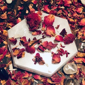 May include: A white hexagonal crystal charging plate with dried rose petals scattered on top. The plate is surrounded by more rose petals on a wooden surface.