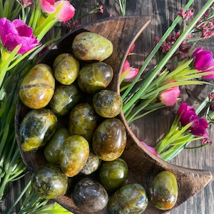 May include: A collection of polished green stones, possibly gemstones, displayed in a wooden crescent-shaped bowl. The stones vary in shade from light to dark green, with some showing brown and yellow hues. Pink and green floral accents surround the bowl.