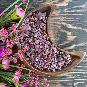 May include: A wooden crescent moon bowl filled with pink and red ruby gemstone chips. The bowl is on a dark wood surface.